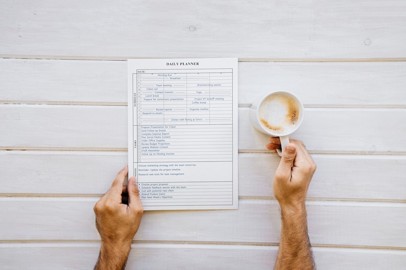 Puede incluir: Un planificador diario con un horario detallado y una lista de tareas es sostenido por una persona. Una taza blanca de caf&eacute; con una capa de espuma se sostiene en la otra mano. El planificador y la taza est&aacute;n sobre una superficie de madera blanca.