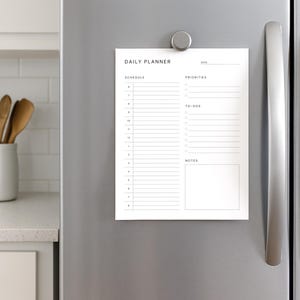 Overhead view of a printed minimalist daily planner page with clean black text on white background, featuring hourly time slots and task list, surrounded by notebooks and pens.