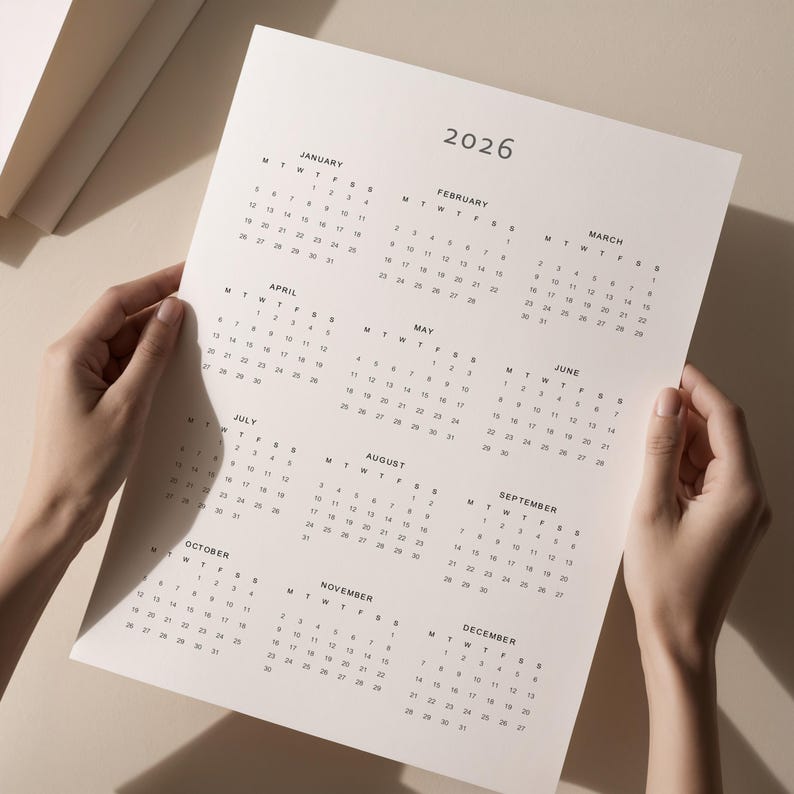 Minimalist 2026 full-year calendar with Monday and Sunday start options, printed on US Letter paper, held by a hand against a plain beige wall background, clean and simple style.