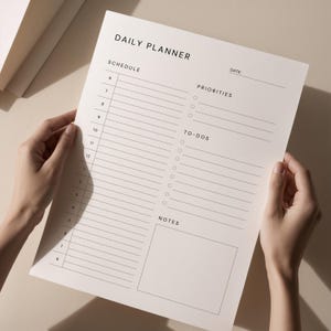 A hand places a printed daily planner sheet into a binder on a neutral workspace, showing neat black typography, clean lines, and organized task list and notes boxes.