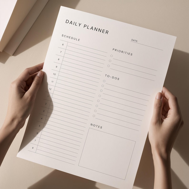 A hand places a printed daily planner sheet into a binder on a neutral workspace, showing neat black typography, clean lines, and organized task list and notes boxes.