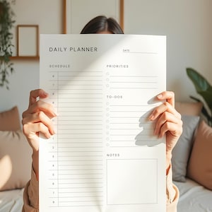 A minimalist daily planner template printed on matte white paper, displaying priorities, notes, and schedule columns, surrounded by stationery on a light wooden desk.