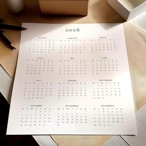 2026 calendar printable PDF shown printed on white paper with full year overview grid, clean grey typography and minimalist layout, held by a hand against a light neutral background setting