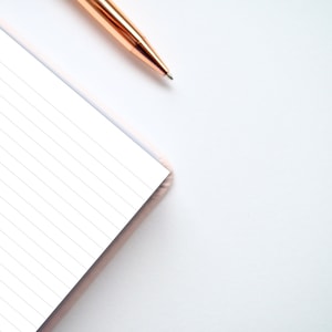 May include: A close-up of a lined notebook with a pink cover and a gold pen lying on a white surface.