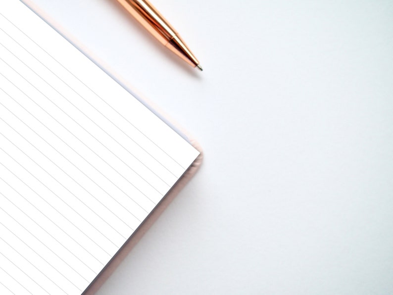 May include: A close-up of a lined notebook with a pink cover and a gold pen lying on a white surface.