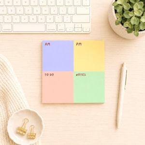 Peut inclure: Un bloc-notes carré coloré divisé en quatre sections : AM (bleu), PM (jaune), TO DO (rose) et NOTES (vert). Un stylo blanc, un clavier, une petite plante dans un pot blanc et des trombones dorés sont également visibles.