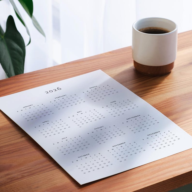 Grey modern font 2026 one-page calendar on white A3 sheet, featuring portrait layout and all twelve months visible, pinned to a white textured wall, photographed in natural daylight.