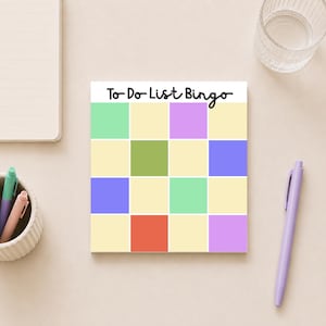 May include: A colorful "To-Do List Bingo" notepad with a grid of squares in pastel colors. The notepad is surrounded by a pen, a glass of water, and other stationery items on a light beige surface.