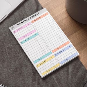 May include: A white monthly budget notepad with sections for income, expenses, bills, savings, and a summary. The notepad has a colorful design with pink, orange, blue, and yellow accents. The text "MONTHLY BUDGET" is at the top.