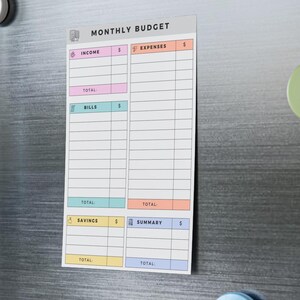 Cute budget tracker with 50 adhesive sheets, structured rows for financial logging, and icons beside each labeled section, placed flat on a light wood surface.
