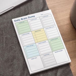 Peut inclure: Bloc-notes blanc "Daily Brain Dump" avec des sections pour les tâches, les rendez-vous et les notes. Le bloc-notes a une mise en page en grille avec des sections codées par couleur en vert, bleu et jaune. Le texte comprend "I need to call", "Appointments" et "Random thoughts".