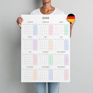 Könnte beinhalten: Ein großer Kalender für 2026 mit monatlichen Layouts in Pastellfarben. Der Kalender wird von einer Person gehalten und zeigt das Jahr oben. Die deutsche Flagge ist in der oberen rechten Ecke sichtbar.