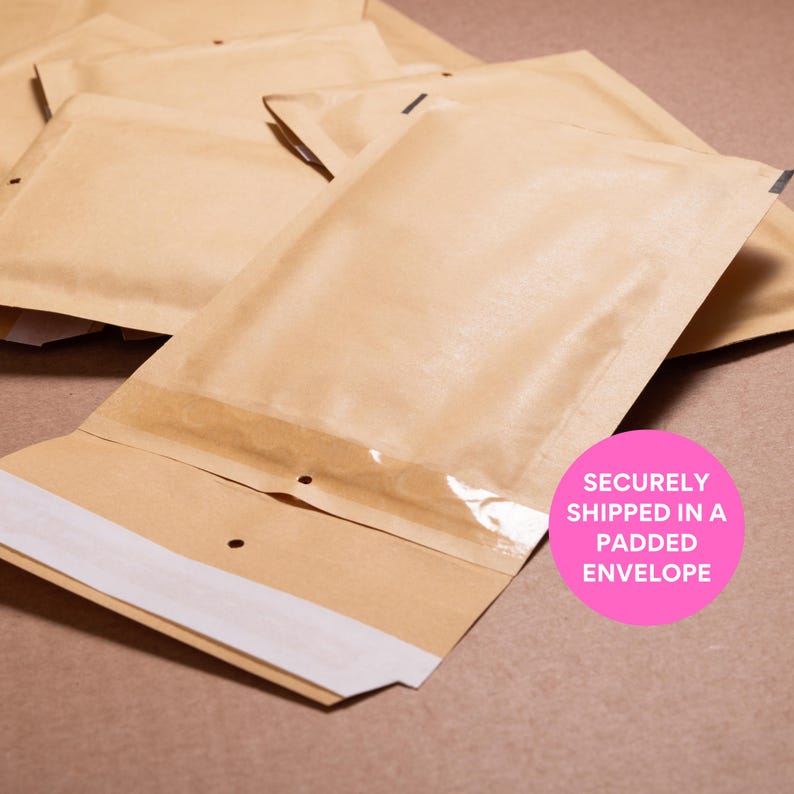 May include: Several brown padded envelopes are stacked on a brown surface. The envelopes have a clear plastic window and a white adhesive strip. A pink circle with the text "SECURELY SHIPPED IN A PADDED ENVELOPE" is in the lower right corner.