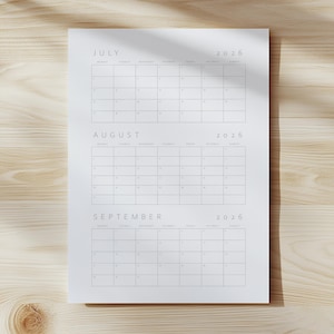 Op de afbeelding: Een witte papieren kalender voor juli, augustus en september 2026. De kalender heeft een minimalistisch ontwerp met een rasterindeling en lichtgrijze tekst. De kalender ligt op een licht houten oppervlak.
