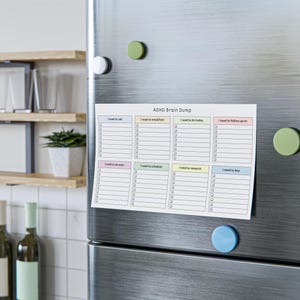 Close-up of a pastel headed ADHD brain dump checklist notepad showing eight sections like “I need to buy” and “I need to schedule” with checkboxes, photographed on a wooden desk surface.