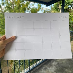 Single month 2026 calendar printed on white paper, landscape orientation, featuring grey minimalist lines and font, lying flat on a neutral medium-light background.