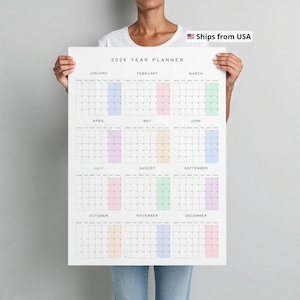 Vertical 2026 wall calendar shown taped to a neutral beige wall, printed on white paper with thin black lines, pastel-colored weekends, and a full year on a single page with Monday start.