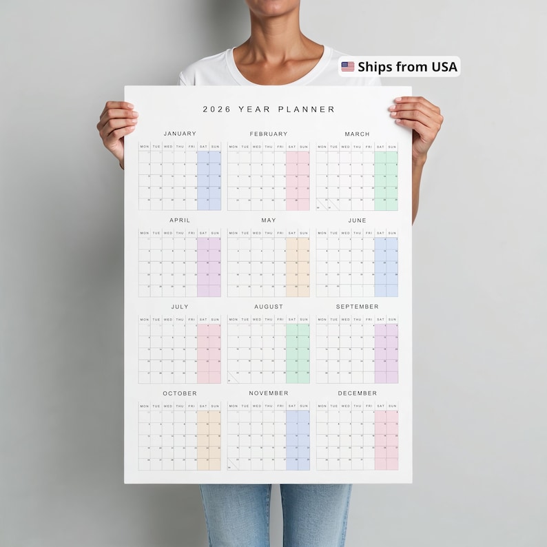 Vertical 2026 wall calendar shown taped to a neutral beige wall, printed on white paper with thin black lines, pastel-colored weekends, and a full year on a single page with Monday start.