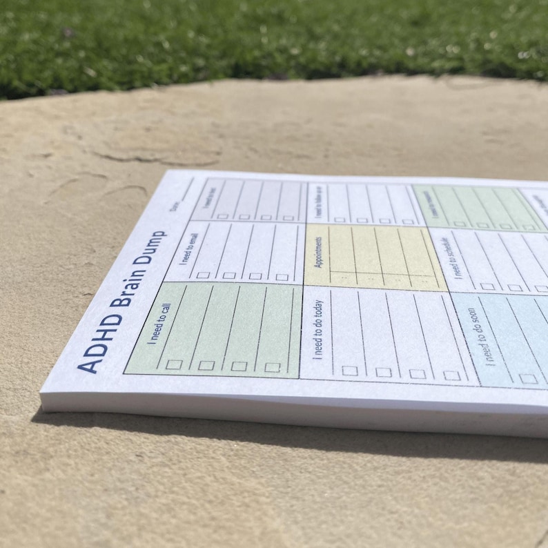 Compact daily brain dump notepad displayed on a large rock outdoors, featuring bold printed labels for planning tasks, appointments, and reminders in a structured format.