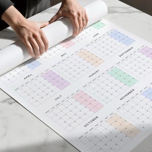 May include: A large, white wall calendar with monthly layouts, featuring pastel-colored boxes for each month. The calendar is being unrolled, revealing the months of April through November. The days of the week are listed across the top.