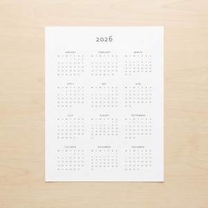 Minimalist 2026 calendar printable shown printed on white paper with portrait layout, full year grid view and clean grey typography, photographed in a modern desk setup environment