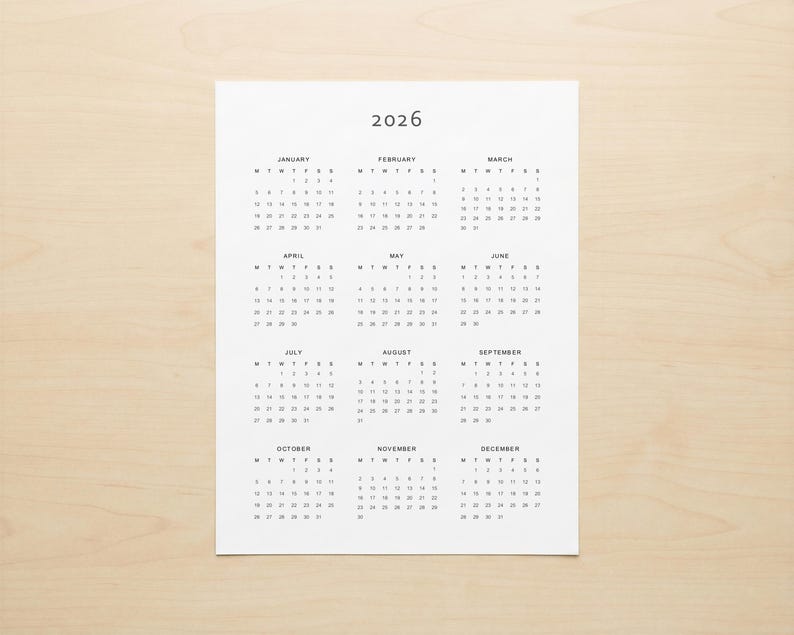Minimalist 2026 calendar printable shown printed on white paper with portrait layout, full year grid view and clean grey typography, photographed in a modern desk setup environment
