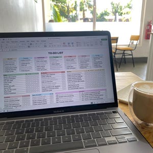 May include: A MacBook Air laptop displaying a detailed to-do list spreadsheet. The screen is filled with rows and columns, outlining tasks for each day of the week. A double-walled glass of coffee sits nearby.