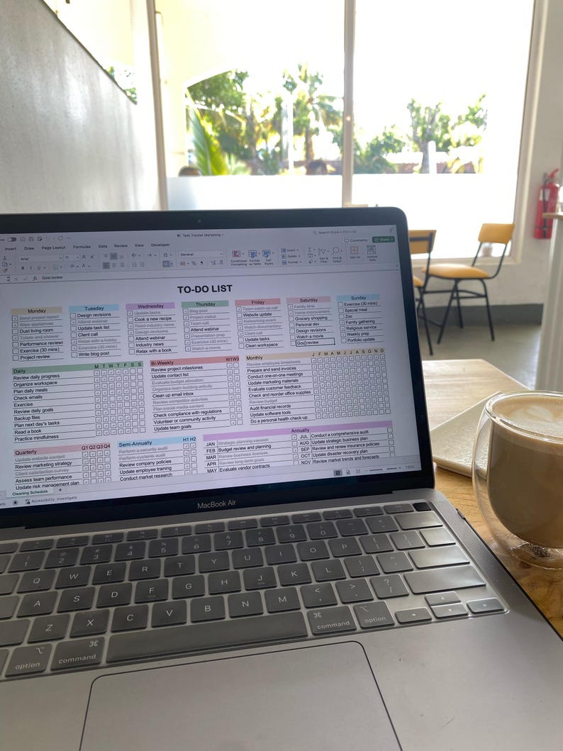 May include: A MacBook Air laptop displaying a detailed to-do list spreadsheet. The screen is filled with rows and columns, outlining tasks for each day of the week. A double-walled glass of coffee sits nearby.