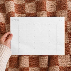 Hand holding an April 2027 printable monthly calendar with a minimalist landscape design against a textured blanket.