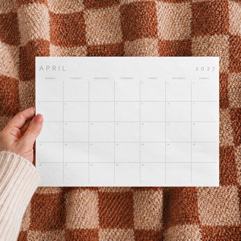 Hand holding an April 2027 printable monthly calendar with a minimalist landscape design against a textured blanket.