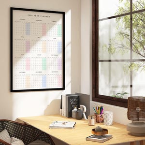 A 2026 calendar poster laid on a desk, showing a clean minimalist layout with Monday start, subtle pastel weekend shades, thin black lines, and full-year view on a single white sheet.