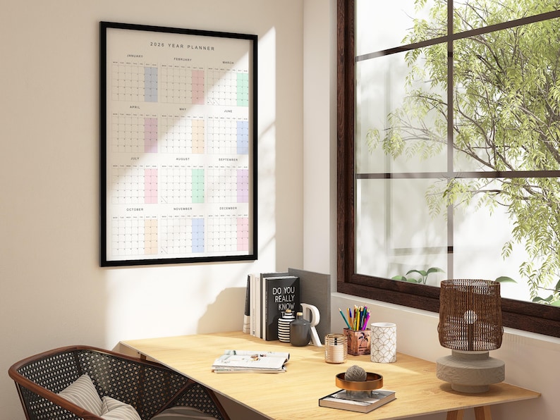 A 2026 calendar poster laid on a desk, showing a clean minimalist layout with Monday start, subtle pastel weekend shades, thin black lines, and full-year view on a single white sheet.