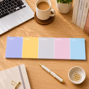 May include: A colorful weekly planner with pastel-colored sections for each day of the week. The days of the week are printed in a light gray font. A white and gold pen, a cup of coffee, and a small plant are also on the wooden desk.