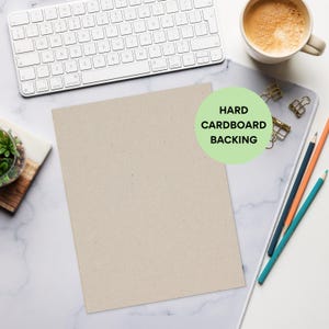 May include: A beige rectangular hard cardboard backing sheet is centered on a white marble surface. A white keyboard, a cup of coffee, pencils, and paper clips are also on the surface. The text "HARD CARDBOARD BACKING" is in a green circle.