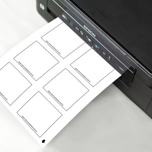 May include: A black and silver printer with a white sheet of paper coming out of the paper tray. The paper has six blank squares with the text "Write Your Adventure Story" in each square.