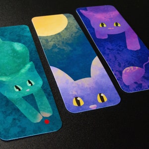 May include: Three watercolor bookmarks featuring cats with different colored fur and backgrounds. The first bookmark features a teal cat with a red dot on a blue background. The second bookmark features a purple cat with yellow eyes on a blue background with a yellow moon. The third bookmark features a purple cat with yellow eyes on a blue background with a purple mouse.