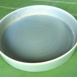 May include: A round, shallow ceramic dish with a smooth, light blue glaze. The interior surface features subtle variations in color, transitioning from a pale sky blue to a deeper, muted blue tone, with a clean, raised rim and a flat base.