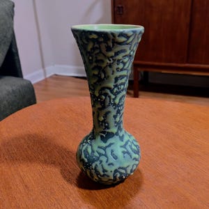 May include: A ceramic vase with a bulbous base and a flared rim. The exterior features a textured, raised pattern in a light lime green color layered over a dark charcoal gray base, creating a distinct contrast across the entire surface.
