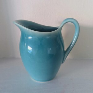 May include: A solid turquoise ceramic creamer pitcher with a smooth, glossy finish. The vessel features a rounded body, a curved pouring spout, and a single arched handle attached to the side. The color is consistent throughout the exterior surface.