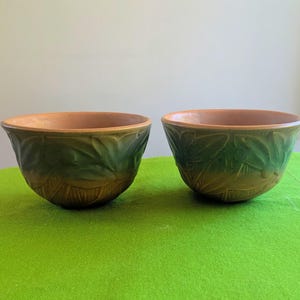 Puede incluir: Dos cuencos de cerámica a juego con un esmalte verde oscuro en el exterior y un patrón de hojas en relieve. El interior de cada cuenco tiene un acabado liso, sin esmaltar, de color terracota, con una forma redondeada y ancha.