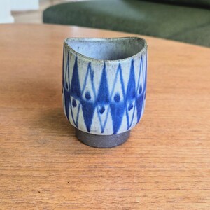 Puede incluir: Una pequeña taza de cerámica de forma cilíndrica con un borde ligeramente irregular. El exterior presenta un patrón repetitivo de diamantes azules pintados sobre una base gris claro, mientras que la base es de material gris oscuro texturizado.
