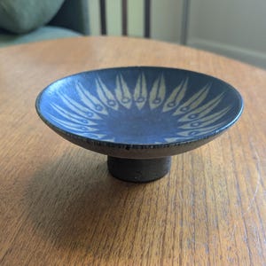 May include: A small, circular ceramic bowl with a pedestal base. The interior features a deep blue glaze with a radiating pattern of light-colored, leaf-like shapes. The exterior of the bowl and the cylindrical base have a dark, textured finish.