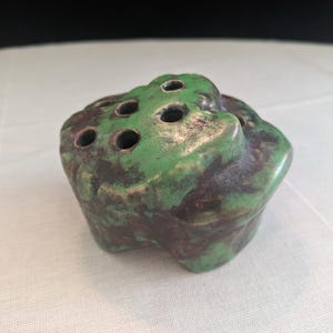 May include: A ceramic flower frog with a mottled green and brown glaze. The frog has a rounded, organic shape with several holes on the top for arranging flowers. The glaze has a textured appearance.