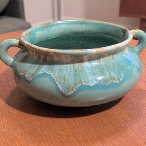 May include: A round ceramic bowl featuring two side handles and a wide opening. The exterior and interior display a glossy glaze in shades of turquoise and teal, with a distinctive drip pattern in a lighter, creamy tan color around the rim.