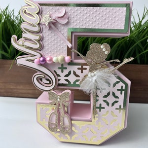 May include: A pink and gold number five cake topper with a ballerina, a wand, and a pearl necklace. The topper says "Viva" on the side.