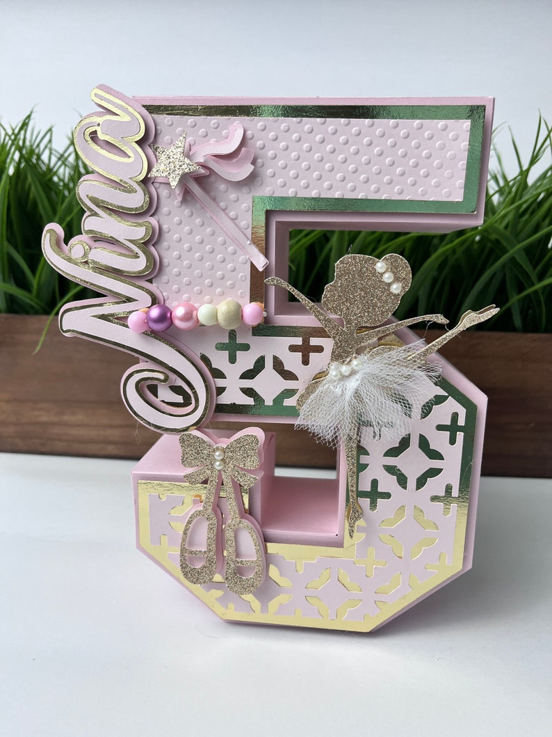 May include: A pink and gold number five cake topper with a ballerina, a wand, and a pearl necklace. The topper says "Viva" on the side.