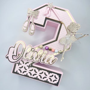 May include: Pink and gold number two cake topper with a ballerina, a bow, and the name Olivia.