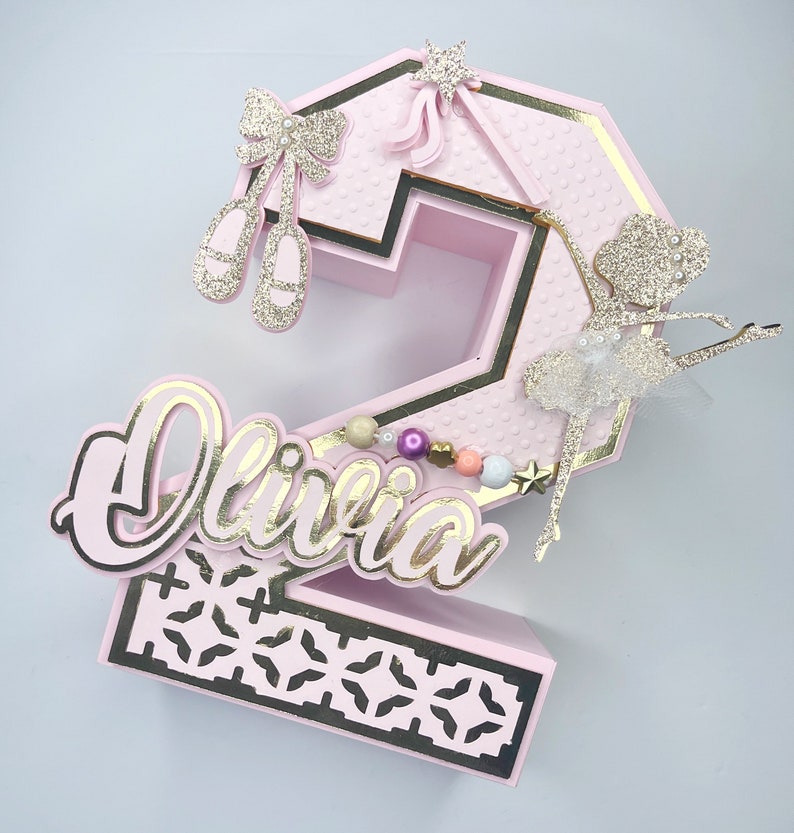 May include: Pink and gold number two cake topper with a ballerina, a bow, and the name Olivia.