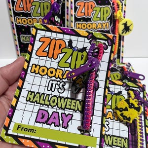 Puede incluir: Una tarjeta con tem&aacute;tica de Halloween con un brazalete de cremallera morado y rosa adjunto. La tarjeta tiene un fondo verde y amarillo con el texto "ZIPZIP HOORAY! IT'S HALLOWEEN DAY" y un espacio para "From:".