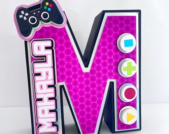 Könnte beinhalten: Ein rosa Buchstabe M mit schwarzem Rand und einem Wabenmuster. Der Buchstabe hat oben links einen schwarzen und grünen Videospiel-Controller und einen weißen Rand mit dem Wort "Happy" in pixelartigen weißen Buchstaben. Der Buchstabe hat auch vier weiße Kreise mit farbigen Knöpfen auf der rechten Seite: ein blaues Quadrat, ein grünes Pluszeichen, ein roter Kreis und ein gelber Play-Button.
